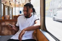 Joven hombre afroamericano positivo en ropa casual navegando en el teléfono móvil mientras está sentado en el tren en Viena Ferrocarril durante el día en la ciudad - foto de stock
