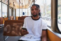 Joven hombre afroamericano positivo en ropa casual navegando en el teléfono móvil mientras está sentado en el tren en Viena Ferrocarril durante el día en la ciudad - foto de stock