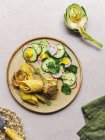 Teller von oben mit Omeletterolle und Rettich-Gurkensalat mit Kräutern in der Nähe von Artischocke und Serviette auf grauem Tisch — Stockfoto