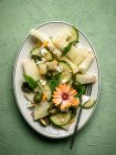 Верхний вид на вкусный салат из дыни с огурцами и оливками подается на тарелке с травами возле солонки и салфетки на зеленом фоне — стоковое фото