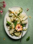 Top view of delicious melon salad with cucumbers and olives served on plate with herbs near salt shaker and napkin on green background — Stock Photo