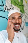 Cheerful Muslim male in traditional clothes smiling and using cellphone while standing near wall on street — Stock Photo