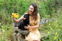 Весела жінка бере самостріл з чорним пухнастим цуценям, використовуючи смартфон і сидячи на дерев'яній лавці в сільській місцевості — стокове фото