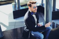 Содержание мужчины в стильной одежде с наушниками и сотовым телефоном, сидящего в городе в солнечный день — стоковое фото