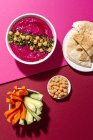 Draufsicht auf appetitliche Rote-Bete-Hummus garniert mit Kichererbsen auf zwei farbigen Hintergrund mit Brot und frischen Möhren-Gurken-Sticks serviert — Stockfoto