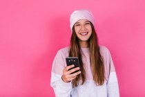 Adolescente femenina feliz con cabello castaño en el pañuelo para la cabeza para el concepto de cáncer navegar por el teléfono celular mirando a la cámara - foto de stock