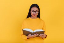 Intelligente asiatico femmina studente in occhiali lettura libro di testo e preparazione per esame su giallo sfondo in studio — Foto stock