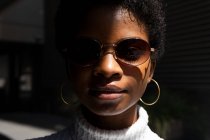 Giovane donna afroamericana in elegante maglione e occhiali da sole guardando la fotocamera mentre in piedi in piena luce solare contro lo sfondo nero — Foto stock