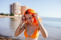 Ritratto di giovane donna rossa sorridente che tocca la testa e guarda la spiaggia in una giornata estiva soleggiata — Foto stock