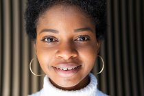 Щаслива стильна молода чорна жінка в комбінованому светрі дивиться на камеру, стоячи біля смугастої стіни будівлі в місті — стокове фото