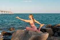 Vue latérale de paisible femme flexible pratiquant le yoga à Eka Pada Rajakapotasana sur le rocher sur la plage — Photo de stock