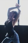 Giovane femmina scalza in jeans con hair bun danza mentre alza lo sguardo sul pavimento con ombre alla luce del sole — Foto stock