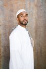 Macho islâmico em roupas brancas autênticas olhando para longe, enquanto de pé contra a parede grungy — Fotografia de Stock