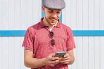 Conteúdo jovem hipster barbudo em camisa de pólo casual e boné navegando telefone celular enquanto está de pé contra a parede branca e azul de madeira sob a luz solar — Fotografia de Stock