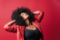 Carefree African American female with Afro hairstyle touching hair with closed eyes on red background in studio — Stock Photo