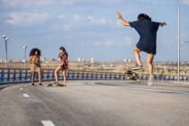 Unrecognizable young woman doing a trick with her long board by a bridge with her companions in the background — Stock Photo