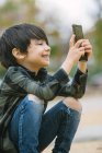Vista lateral do menino adorável em roupas casuais navegando celular enquanto sentado no skate na travessia de pedestres na cidade — Fotografia de Stock