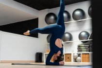 Vista lateral de cuerpo completo de la hembra en forma en ropa deportiva haciendo la postura apoyada de la variación del headstand mientras que practica yoga en estudio de la aptitud - foto de stock