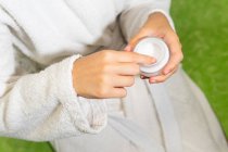Top view of crop young female in white bathrobe sitting on bed and taking cream from jar during skin care routine at home — Stock Photo