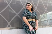 Счастливая молодая пышная женщина в стильном орнаментальном платье и смеясь стоя у стены современного здания — стоковое фото