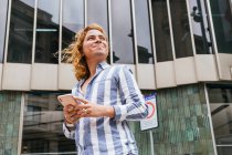 Low angle of tranquil happy male with long hair surfing Internet on mobile phone while standing in city against glass building and looking away — Stock Photo