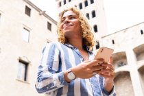 Low angle of tranquil happy male with long hair surfing Internet on mobile phone while standing in city against building and looking away — Stock Photo