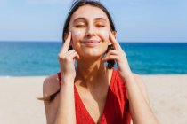 Donna sorridente con gli occhi chiusi che applica lozione abbronzante sul viso nella giornata di sole in estate in spiaggia — Foto stock