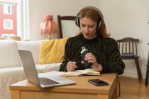 Receptora de radio femenina enfocada con micrófono y auriculares escribiendo en bloc de notas mientras se prepara para grabar podcast en casa - foto de stock