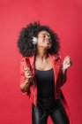 Délicieuse femme afro-américaine écoutant de la musique dans un casque et utilisant un téléphone portable tout en dansant sur fond rouge en studio — Photo de stock