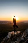 Молода йогиня практикує йогу на скелі в горах зі світлом сходу сонця, вид ззаду з однією ногою і піднятими руками — стокове фото