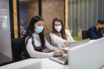Multiracial female colleagues wearing masks sitting at table with laptop and discussing business project while working in coworking space — Stock Photo