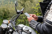 Side view of male biker in trendy leather jacket with rivets and protective helmet browsing phone while sitting on motorcycle parked near lush green forest — Stock Photo