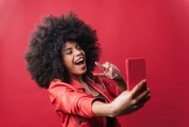 Sorridente donna afro-americana con acconciatura afro scattare autoritratto sul telefono cellulare su sfondo rosso in studio — Foto stock