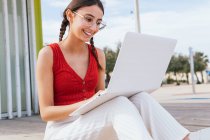 Contenu femme freelance assis sur le bord de la mer et tapant sur ordinateur portable tout en travaillant à distance sur le projet en été — Photo de stock