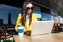 Asiatische Geschäftsfrau mit gelbem Mantel sitzt an einem Tisch und trinkt Kaffee mit ihrem Smartphone und Laptop — Stockfoto