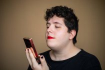 Женский андрогинный мужчина с вьющимися волосами наносит помаду на губы, делая макияж — стоковое фото
