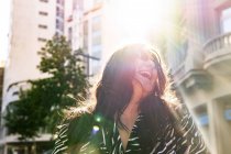 Baixo ângulo de alegre jovem fêmea em roupa elegante e brincos tremendo cabelo à luz do sol na rua urbana — Fotografia de Stock