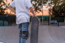 Crop Teenage boy in protective gear standing with skateboard in skate park and looking away — Stock Photo