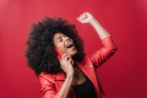 Emocionada mujer afroamericana con el puño cerrado haciendo una llamada en el teléfono móvil y celebrando el éxito en el fondo rojo - foto de stock