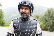 Positive middle aged bearded Hispanic male rider in trendy leather jacket with rivets and black helmet smiling and looking away while standing on road near lush green trees in nature — Stock Photo