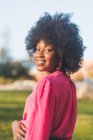 Vue latérale de charmante femme afro-américaine avec les cheveux bouclés souriant à la caméra tout en se tenant debout sur une journée ensoleillée dans le parc — Photo de stock