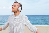 Безтурботний молодий бородатий хлопець у стильній повсякденній сорочці слухає музику через бездротові навушники і насолоджується свіжим вітром, проводячи літній день на піщаному пляжі біля моря — стокове фото