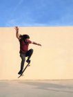 Skateboarder masculin avec des cheveux ondulés effectuant tour sur skateboard tout en sautant par-dessus passerelle et en regardant vers le bas sur la journée ensoleillée avec ciel bleu — Photo de stock