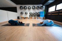 Vista lateral de cuerpo completo de mujeres delgadas y tranquilas que hacen pose Extended Childs mientras estiran el cuerpo y se relajan durante la clase de yoga juntos en el gimnasio - foto de stock