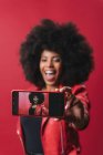 Посмішка афроамериканської жінки з африканською зачіскою робить автопортрет на мобільному телефоні на червоному тлі в студії — стокове фото