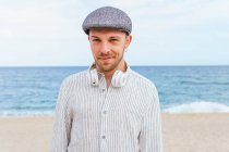 Young bearded male in trendy casual clothes and cap standing on sandy beach towards waving sea while spending summer holidays on seashore — Stock Photo