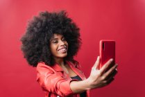 Femme afro-américaine souriante avec coiffure afro prenant autoportrait sur téléphone portable sur fond rouge en studio — Photo de stock