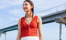 Low angle of young female in stylish glasses and with pigtails standing on background of bridge in city and looking away — Stock Photo