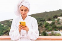 Jovem do sexo feminino em roupão de banho e toalha sorrindo e navegando telefone celular enquanto descansa na varanda após o chuveiro — Fotografia de Stock