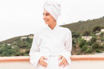 Optimistische junge Frau in Bademantel und Handtuch lächelt und schaut weg, während sie sich bei der Hautpflege am Wochenende auf dem Balkon entspannt — Stockfoto